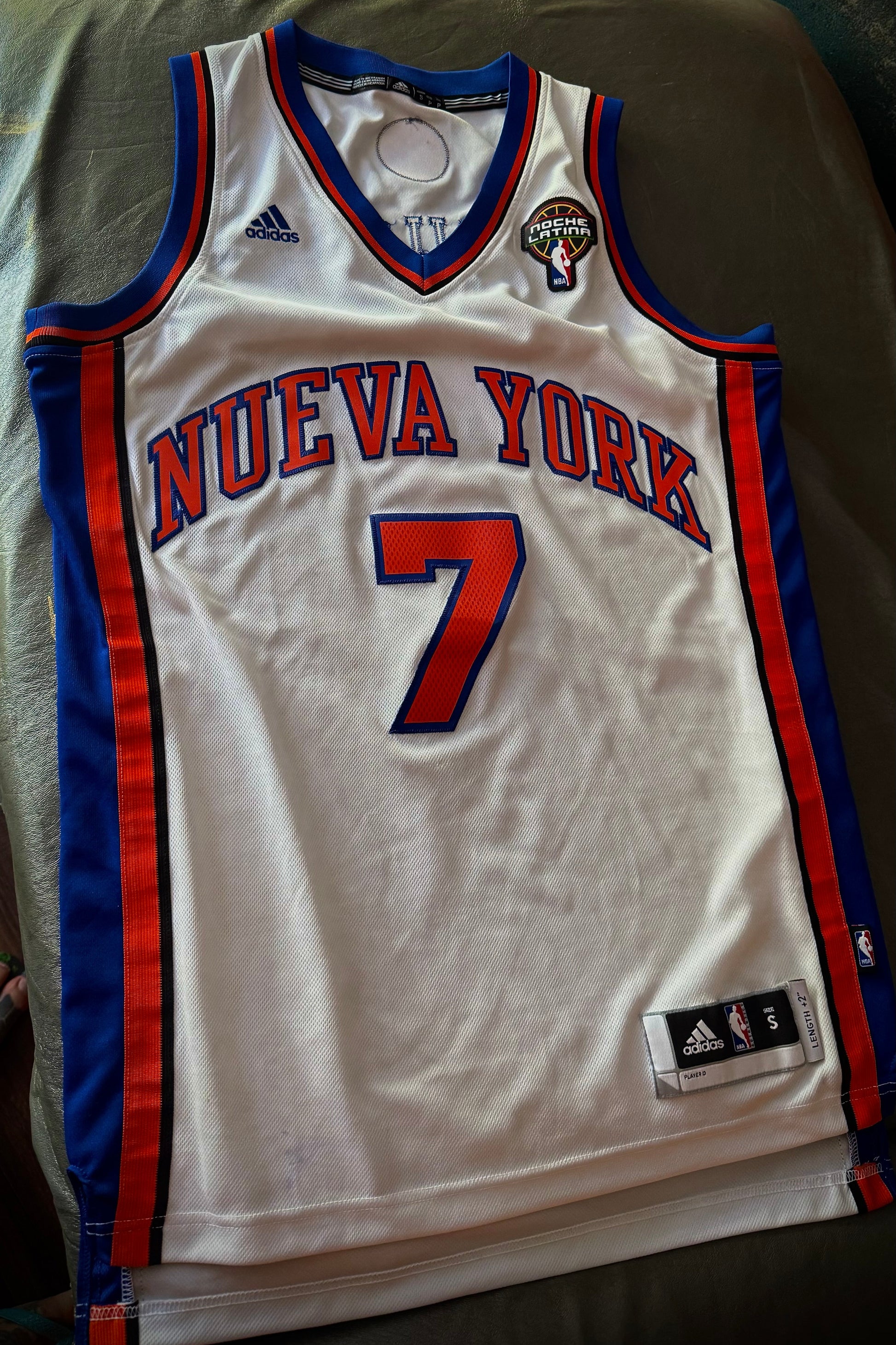 White basketball jersey with red and blue accents, featuring the number 7 and 'Nueva York' text, on a green surface.
