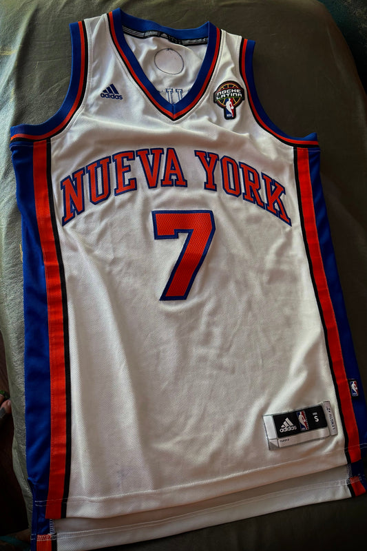 White basketball jersey with red and blue accents, featuring the number 7 and 'Nueva York' text, on a green surface.