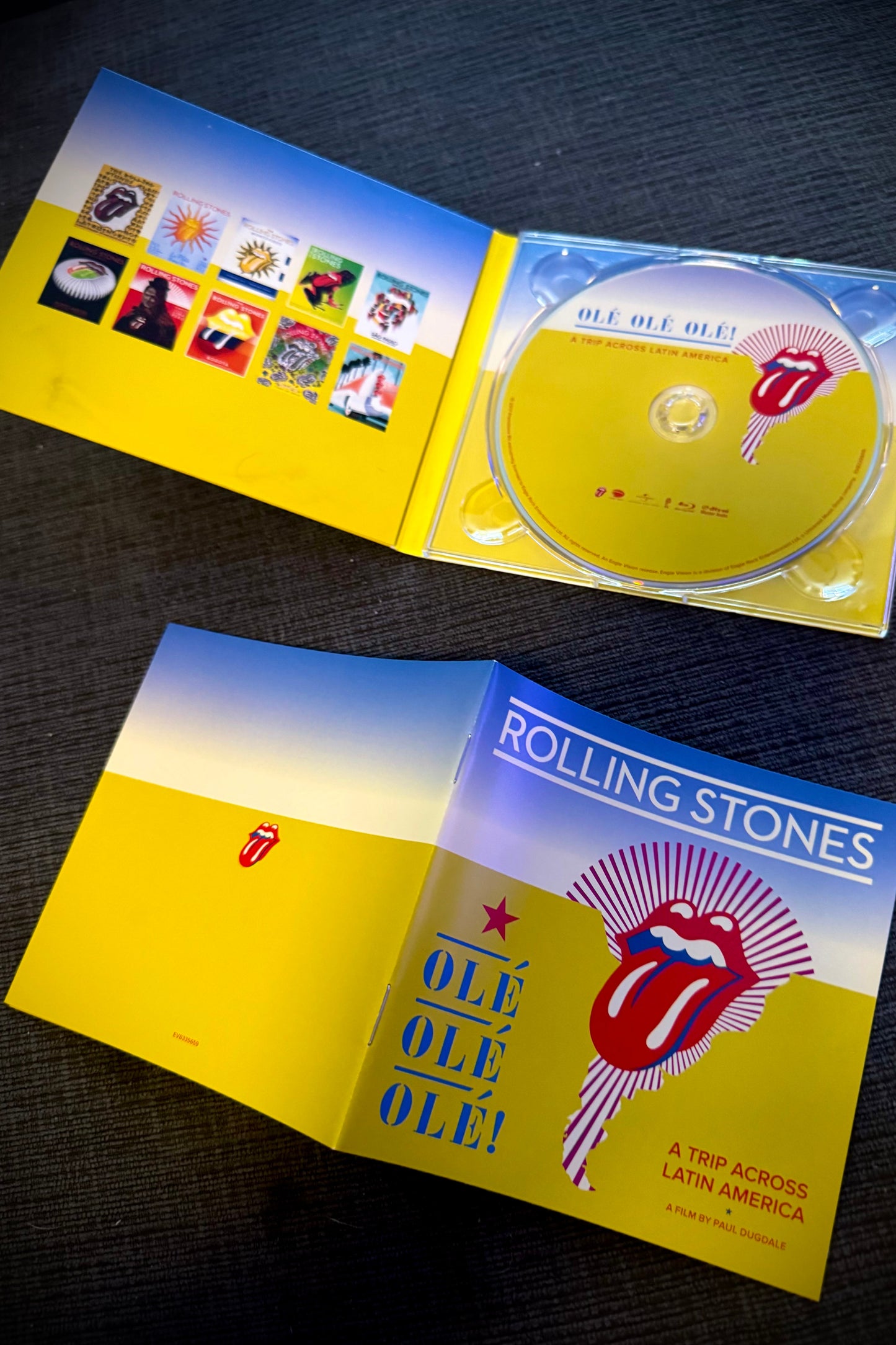 The image shows the Blu-Ray DVD of The Rolling Stones’ Olé Olé Olé!: A Trip Across Latin America. The DVD case is open, displaying the DVD with a red cover and a graphic design, and the inner cover with additional content and images related to the band.