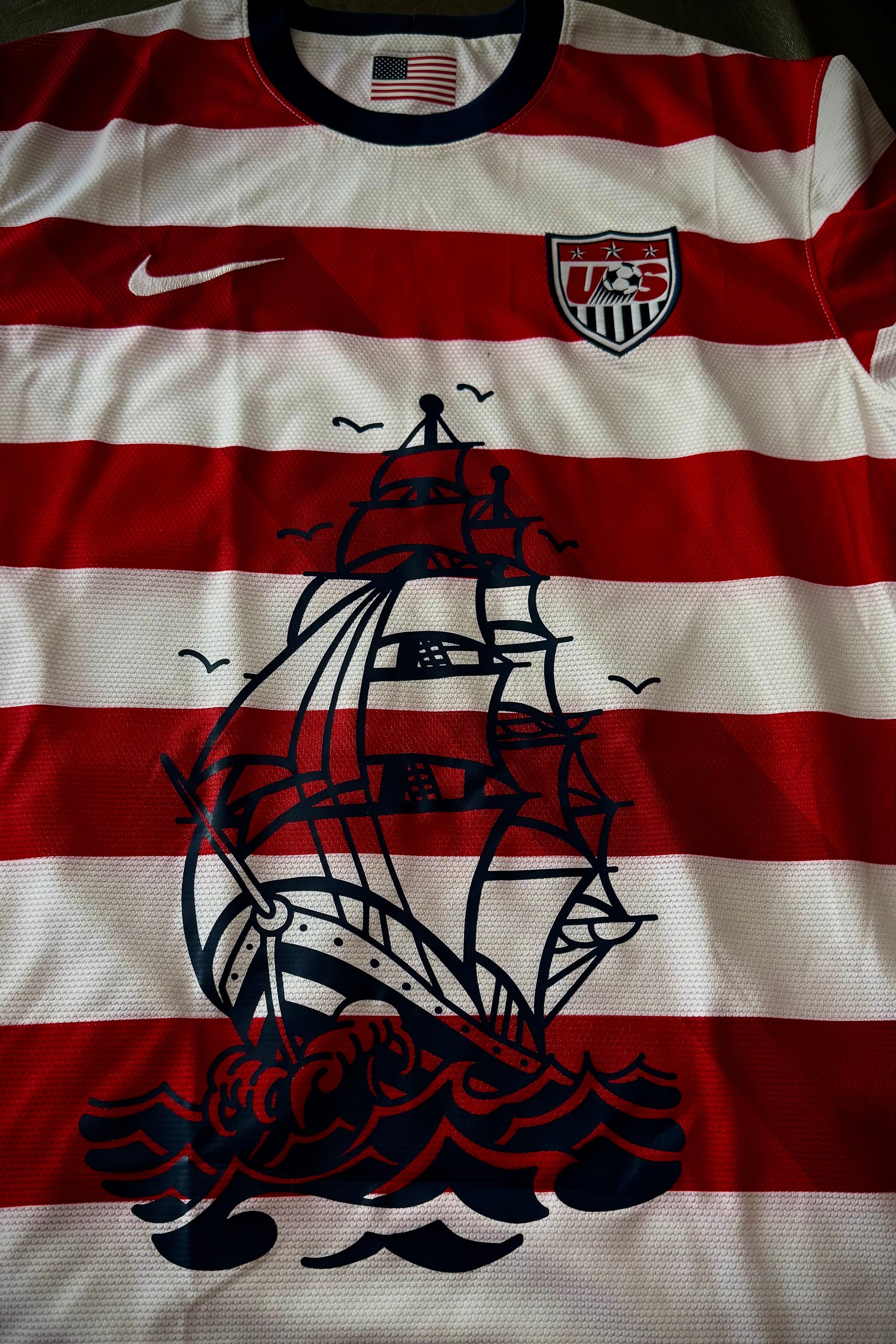 Red and white striped shirt with black ship design and U.S. flag on a dark background